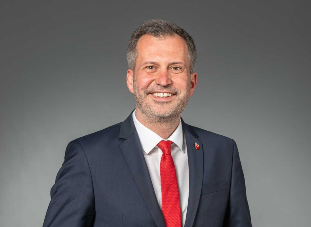 Oberbürgermeister Tobias Schick in einem dunklen Anzug mit roter Krawatte und lächelndem Gesicht vor grauem Hintergrund.